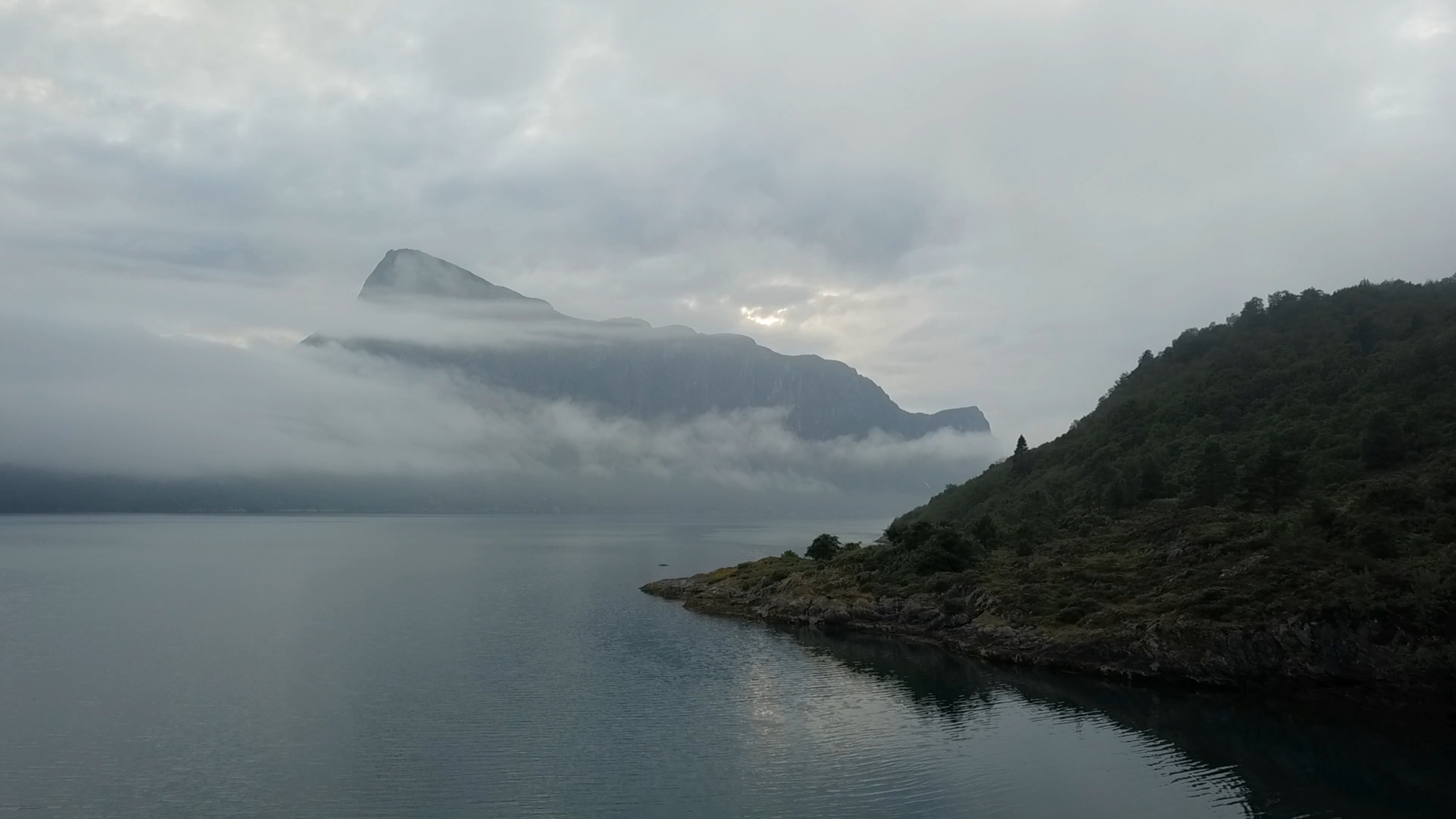 A mountain over a fjord in Norway emerges from a veil of cloud and mist.