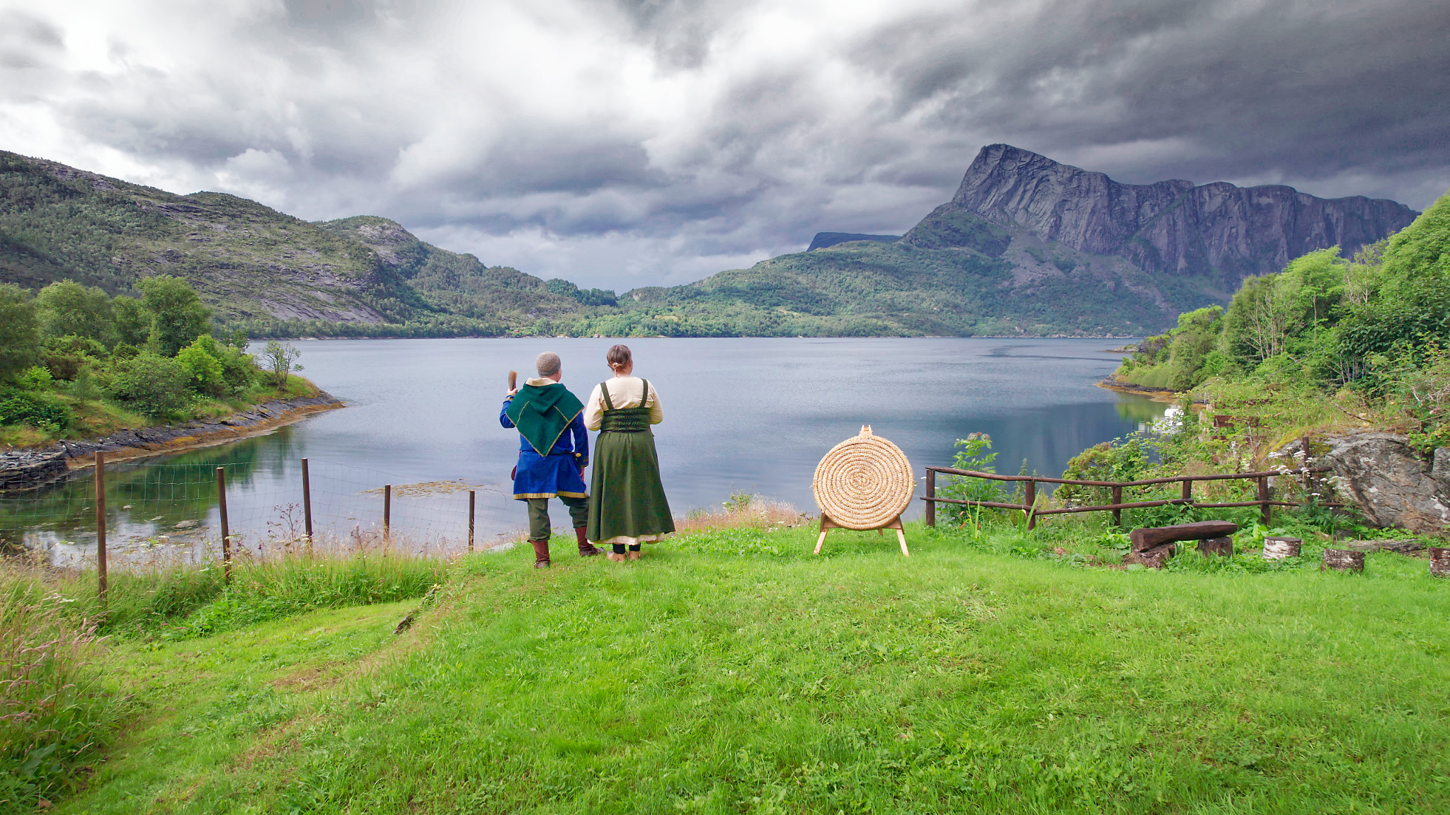 A couple dressed in Viking clothes look out over a Norwegian fjord to the mountain in the distance.